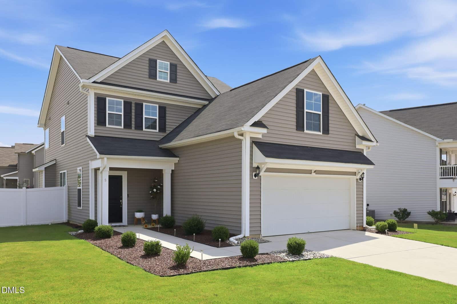 Two-story suburban house with gray siding, white trim, and a double garage. Well-maintained green lawn, small shrubs, and a clear blue sky overhead.