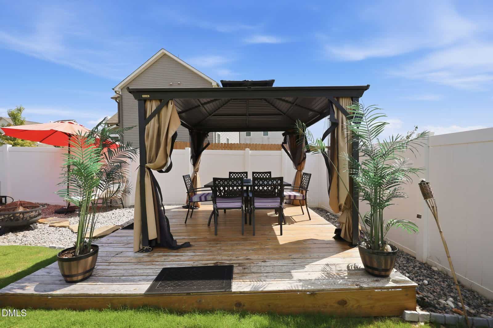 A backyard wooden deck with a black metal gazebo, a dining table and chairs, potted plants, and tiki torches, surrounded by a white privacy fence and a grassy lawn under a blue sky.
