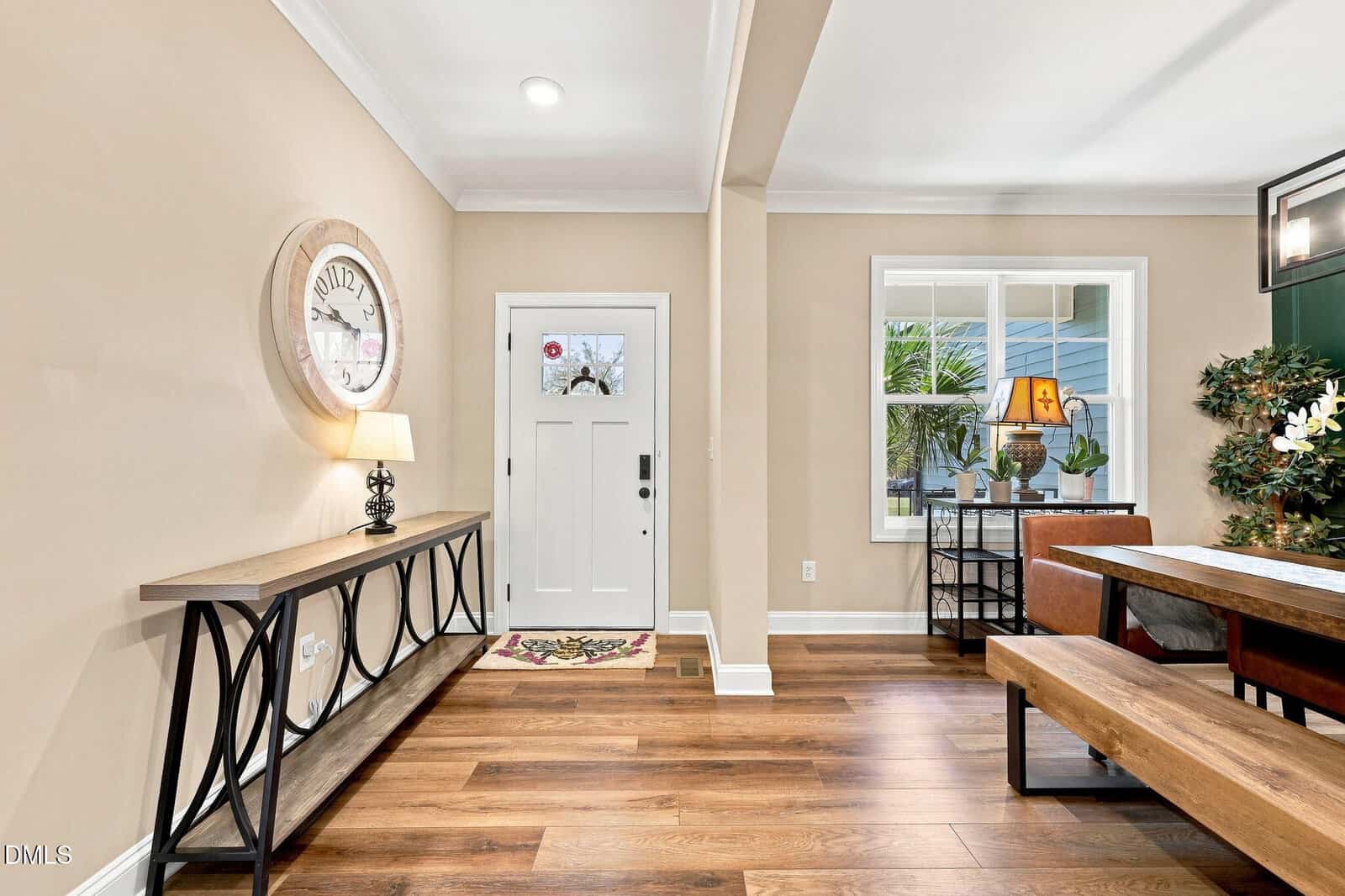 Bright entryway with a white front door, wall clock, console table, and lamp on hardwood floors; adjacent dining area with a wooden table, plants, and large window showing outdoor greenery.