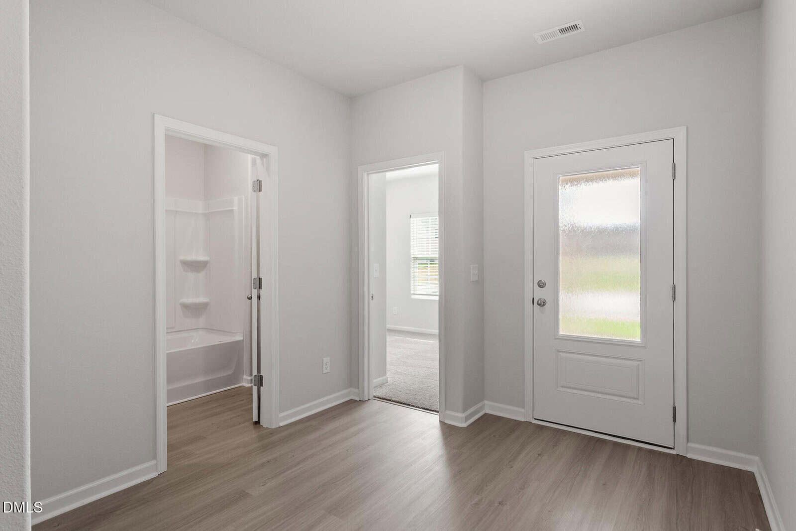 A bright entryway with light wood flooring, a white door with frosted glass, a doorway to a carpeted room, and an open bathroom with a tub and shelves. Walls are white, and natural light fills the space.