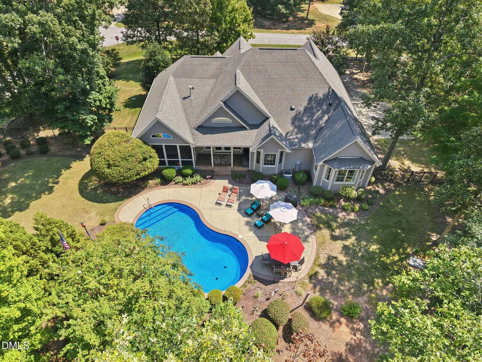 Aerial view of a house with a gray roof, backyard patio, outdoor furniture, umbrellas, and a curved swimming pool surrounded by trees and landscaping on a sunny day.