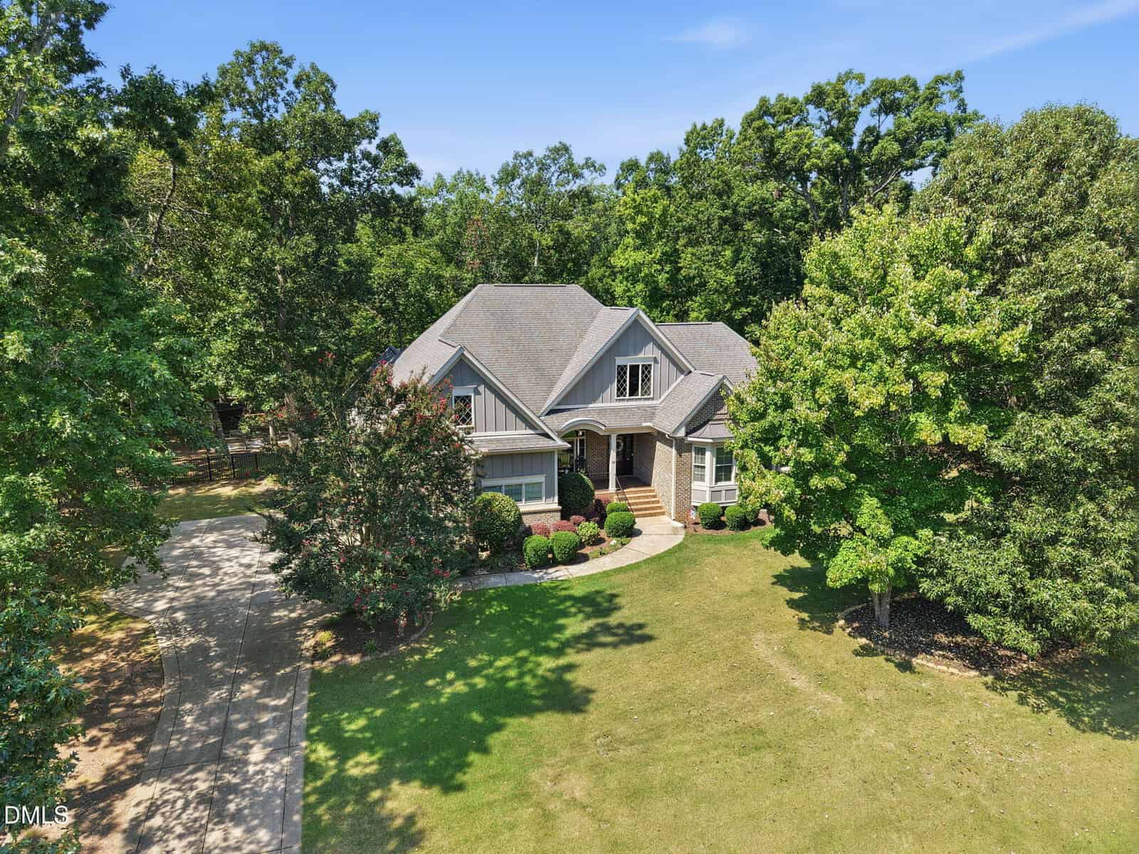 A large gray house with gabled roofs and brick accents sits in the center of a green lawn, surrounded by trees and a curved driveway on a sunny day.