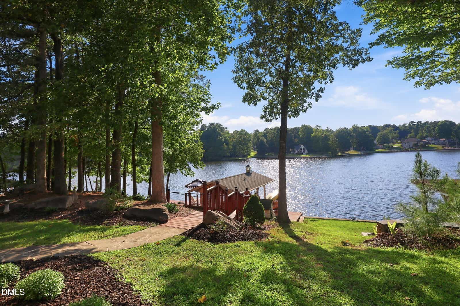 A lakeside scene with a wooden gazebo by the water, surrounded by lush green grass, tall trees, and a clear blue sky reflecting on the lake. A stone path leads down to the gazebo from the foreground.