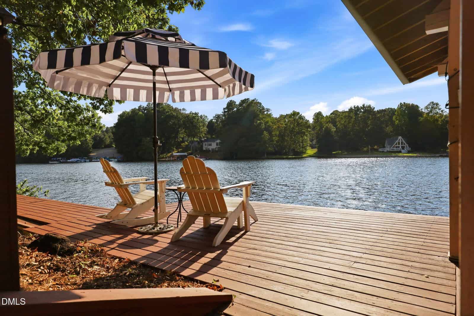 Two wooden chairs and a small table sit on a lakeside deck under a black-and-white striped umbrella, overlooking calm water and trees under a blue sky.