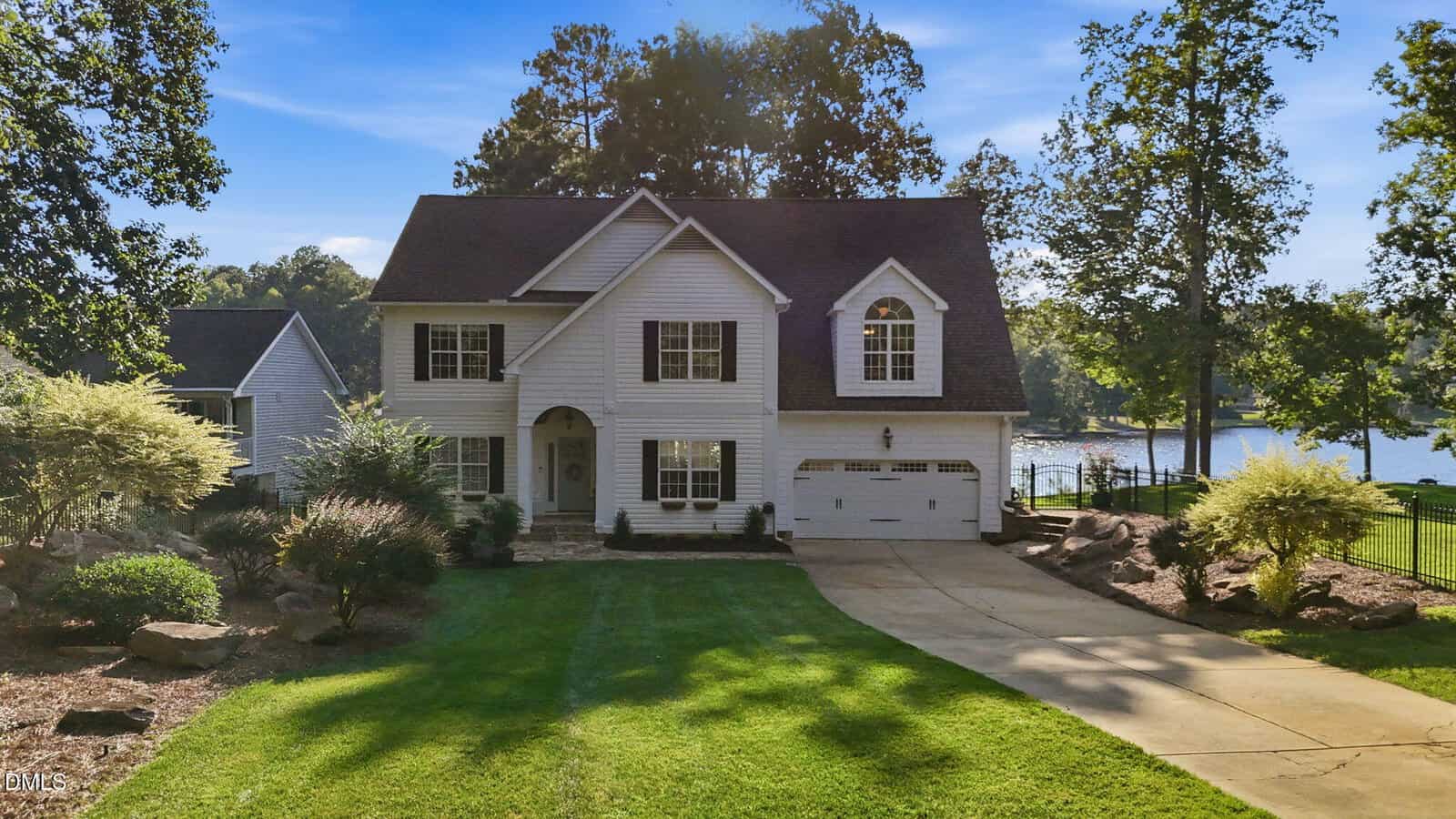 Two-story white house with attached garage, large front lawn, and trees. A paved driveway leads to the house at 1337 Sagamore Drive. A lake and more trees are visible in the background under a clear blue sky, reflecting the peaceful charm of Louisburg.