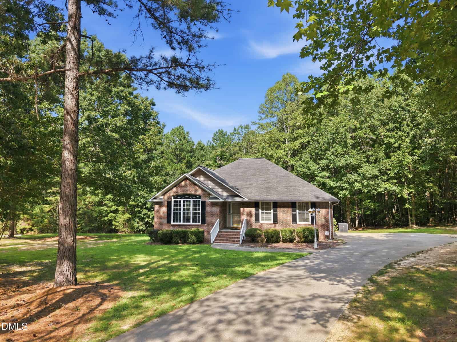 Single-story brick house with large windows, surrounded by trees and greenery at 606 Dabney Woods Lane. A curved driveway leads to the entrance, and the home sits in a peaceful, wooded area of Henderson under a clear blue sky.