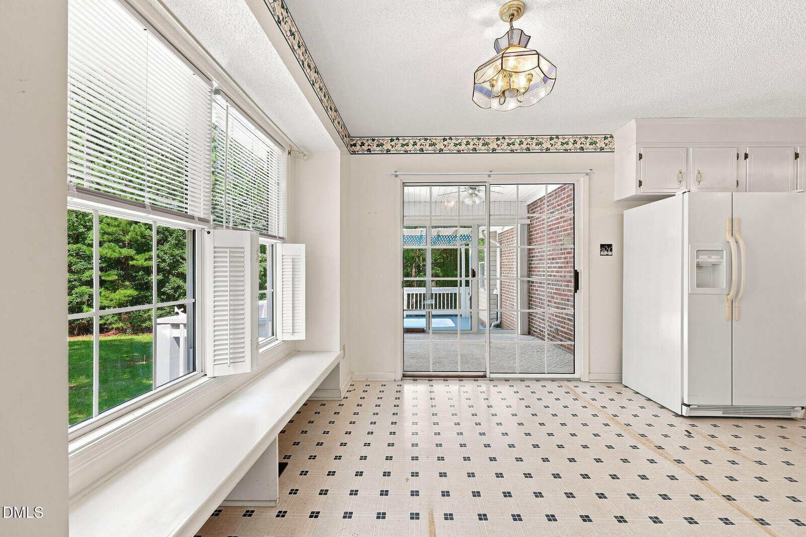 Bright kitchen with patterned floor, large windows, white benches under the windows, sliding glass doors leading to a sunroom, a white refrigerator, and floral wallpaper border near the ceiling.