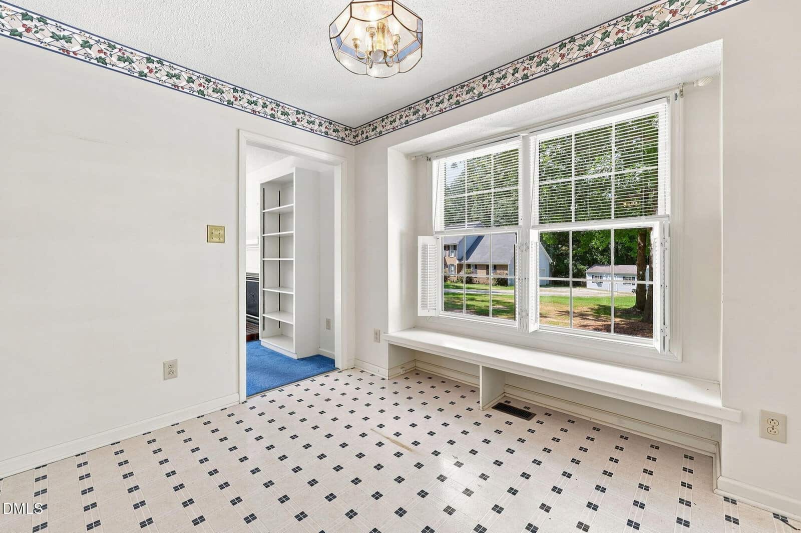 Bright room with patterned tile floor, white walls, a floral border near the ceiling, a large bay window with a bench, and a view into an adjacent room with blue carpet and built-in shelves.