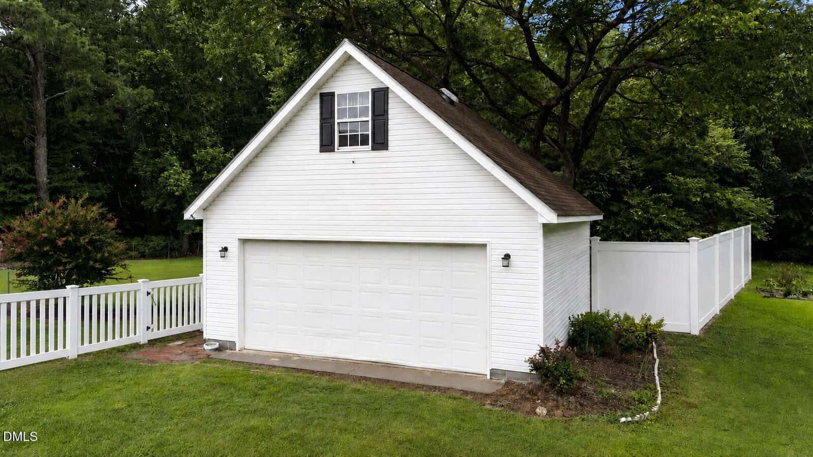 A white detached garage with a gable roof, black shutters, and a double garage door, surrounded by green grass, a white fence, and trees in the background.