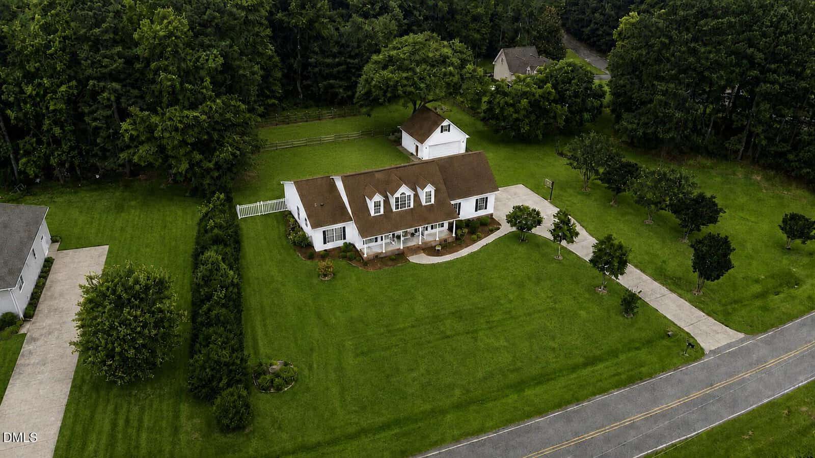 Aerial view of a white house with a brown roof, a detached garage, curved driveway, and large green lawn, surrounded by trees and bordered by a road.