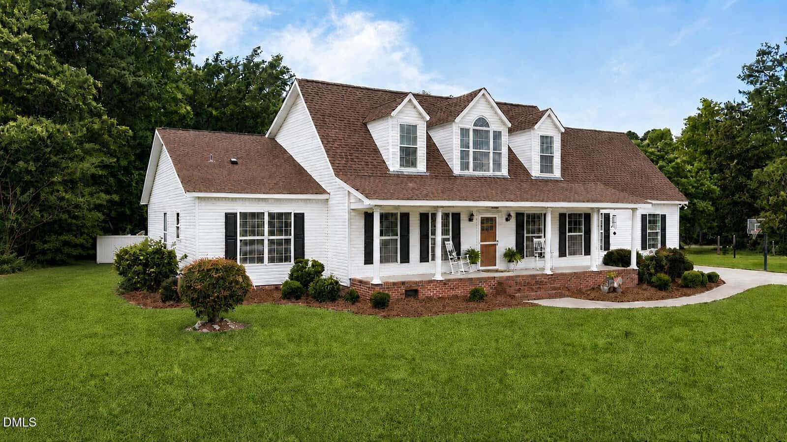 A two-story white house with a brown roof, dormer windows, covered front porch, and a spacious green lawn surrounded by trees under a blue sky sits gracefully at 541 Mallie Pearce Road in Zebulon.