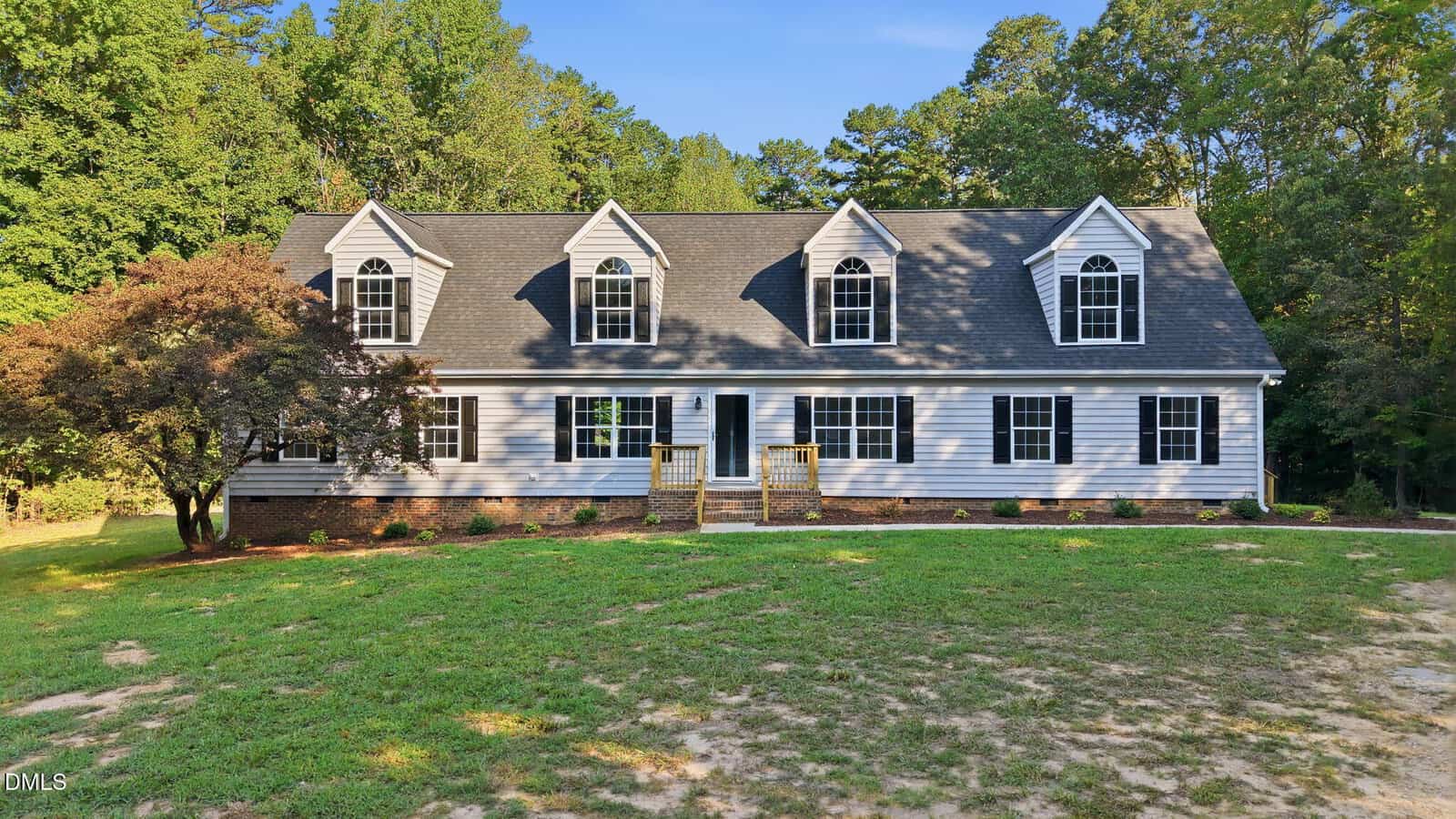 Nestled at 308 Romeo Court in Henderson, this two-story gray house features black shutters, a brick foundation, three dormer windows, and a charming front porch, all surrounded by trees and a lush green lawn under a clear blue sky.