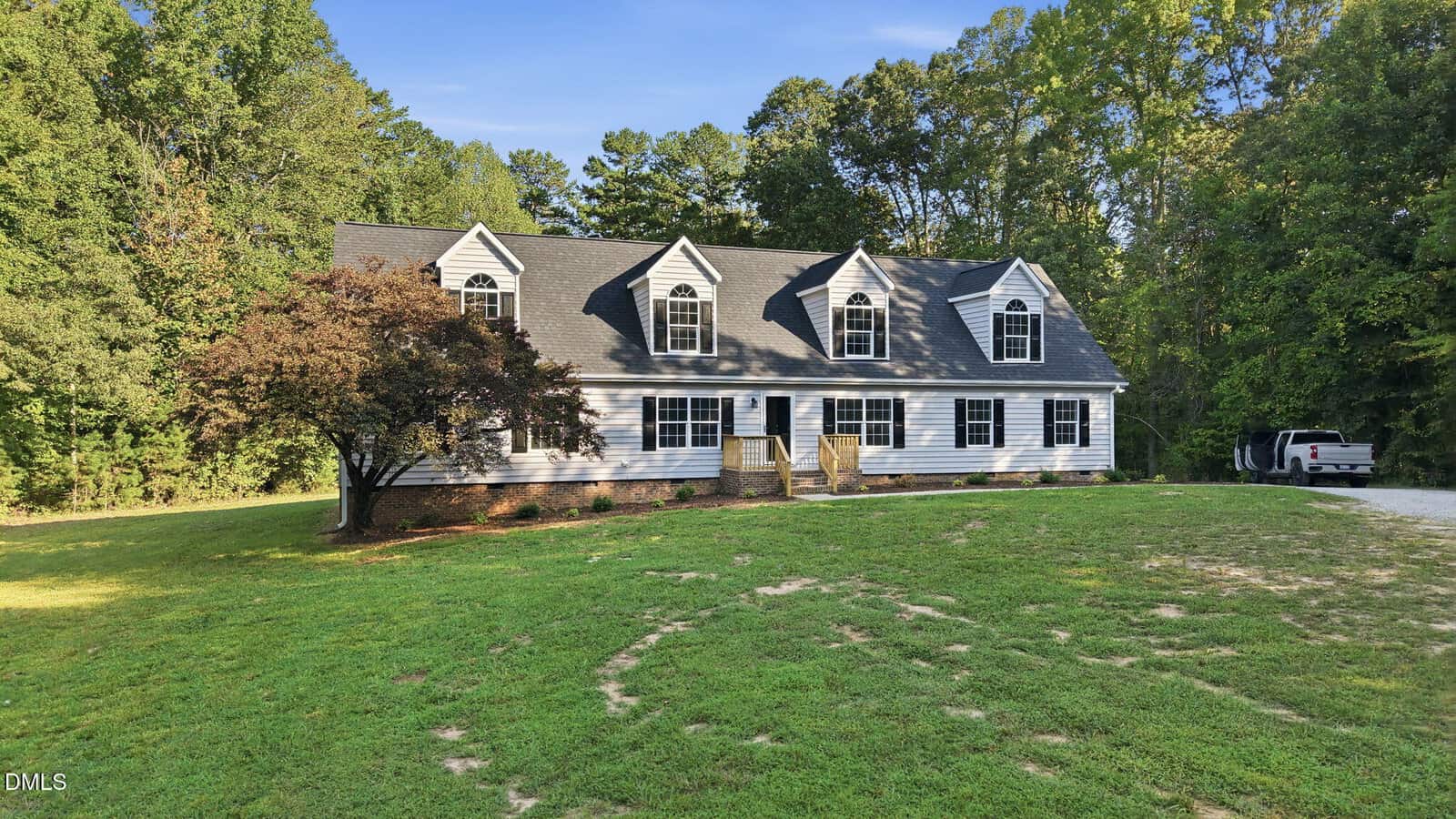 A two-story gray house with dormer windows, black shutters, and a front porch sits on a large grassy lawn, surrounded by trees with a driveway and a parked white vehicle to the right.