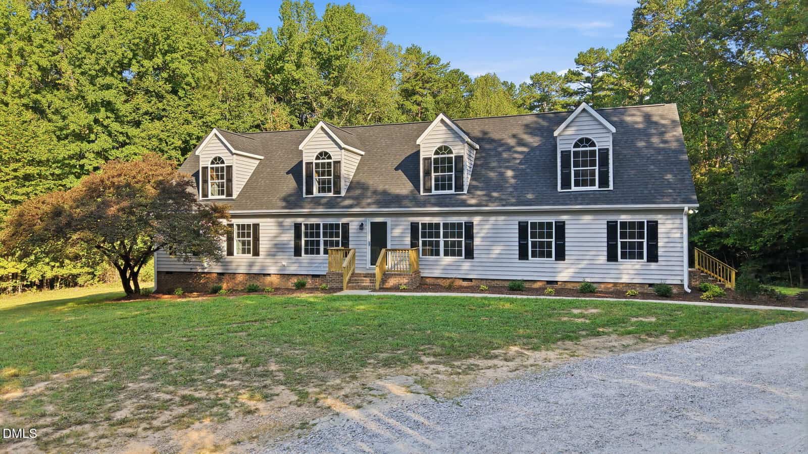 A large white house with black shutters, three dormer windows, and a front porch sits on a spacious lot with trees and a gravel driveway under a blue sky.
