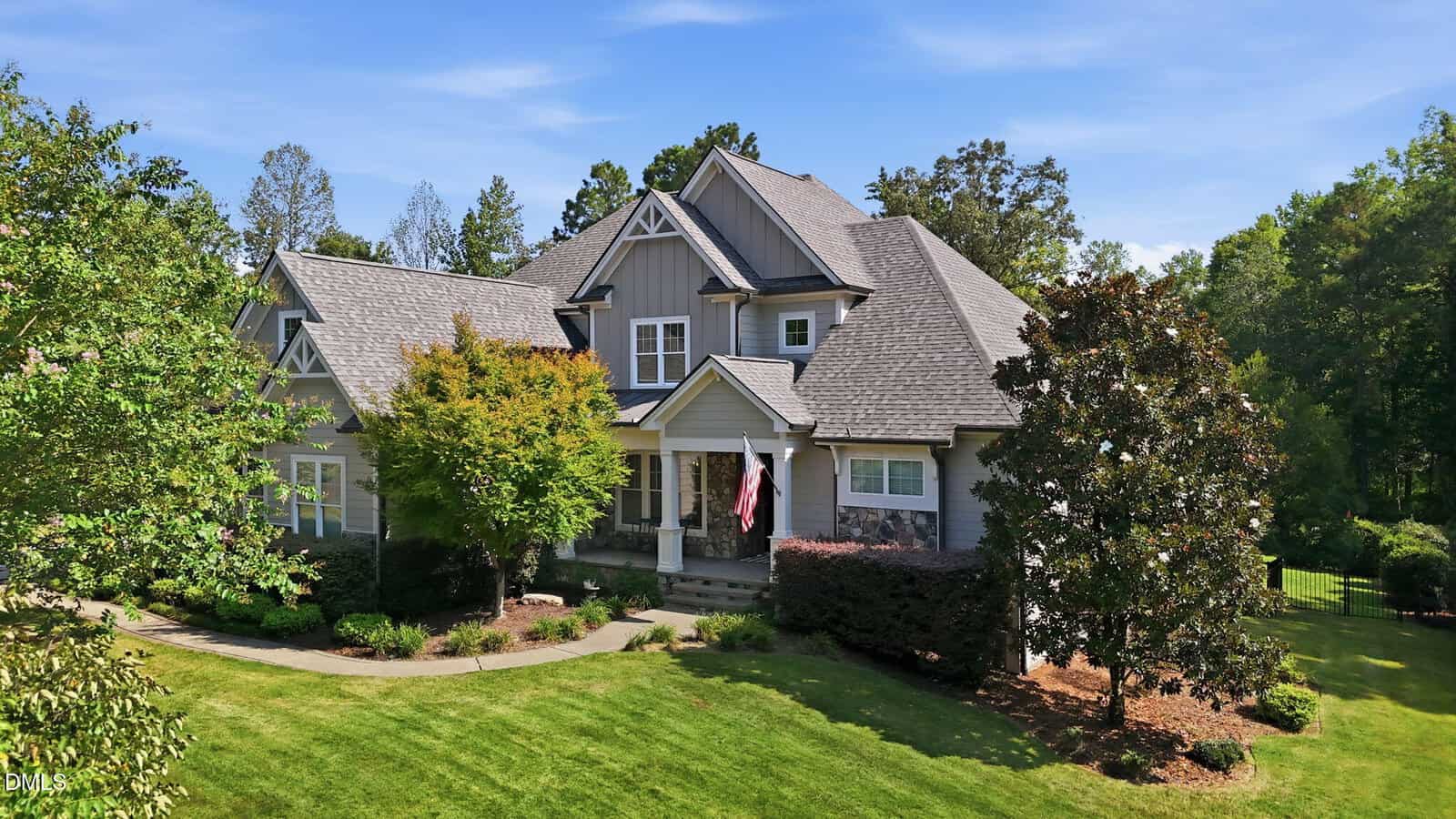 Two-story gray house with gabled roof, front porch, and American flag, nestled at 270 Rivers Edge Drive in Youngsville. Surrounded by a green lawn, mature trees, and landscaped shrubs under a blue sky.