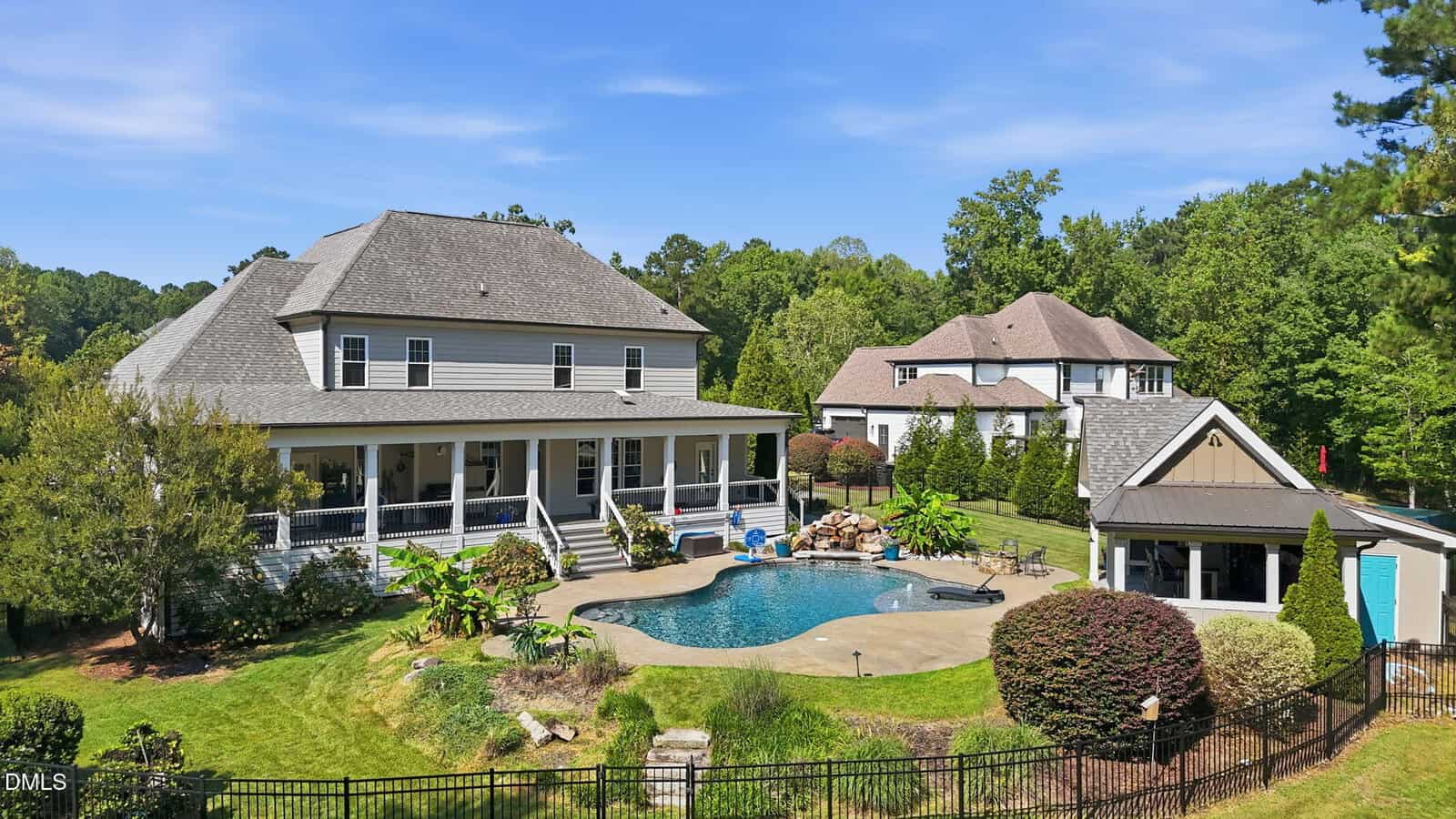Large two-story house with a covered porch, a backyard swimming pool, landscaped greenery, a pool house, and another house nearby, all under a clear blue sky.