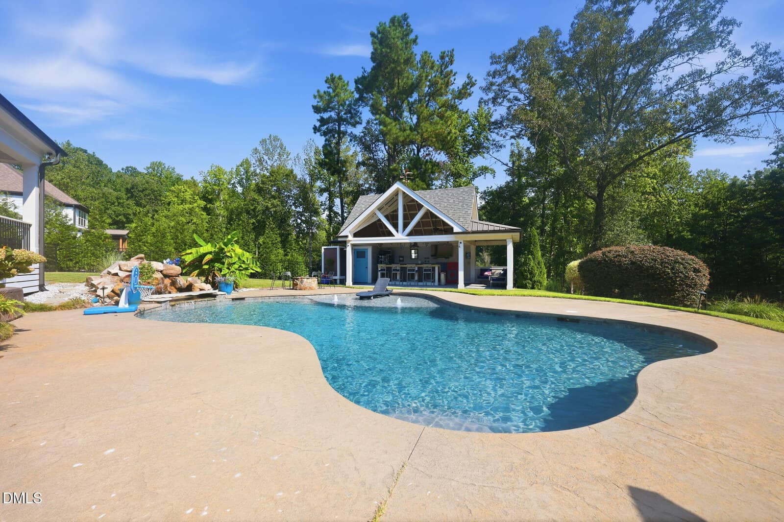 A backyard pool with clear water, a slide, and a waterfall feature, surrounded by trees. There is a pool house with open sides and outdoor seating in the background.