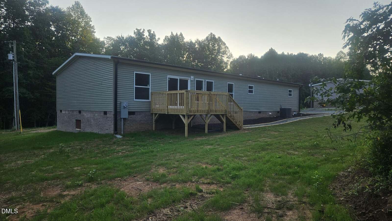A single-story manufactured home with beige siding, black-framed windows, a small wooden deck with stairs, and a grassy yard, surrounded by trees in the background.