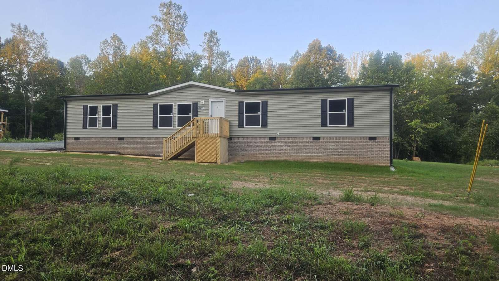 A beige manufactured home with a brick foundation and small wooden steps sits on a grassy lot surrounded by trees under a clear sky.