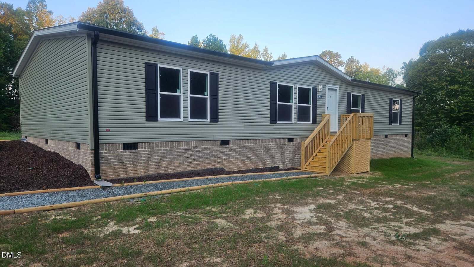Single-story, gray manufactured home with white trim, black shutters, and a brick foundation located at 7635 Sam Hall Road B, Oxford. Wooden steps lead to the front door. The yard is partly grass and bordered by trees under a clear sky.