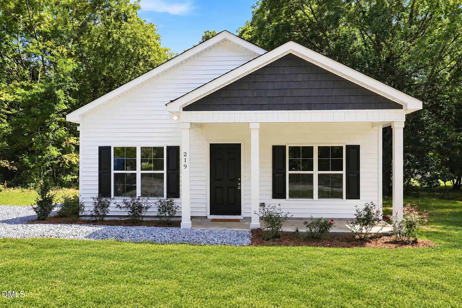 Charming small white single-story house with black shutters and front door at 219 Alexander Avenue in Oxford. Enjoy a covered porch, gravel path, and green lawn surrounded by trees under a bright blue sky.