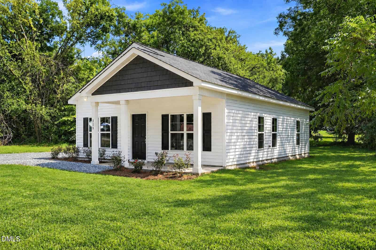 A small white house with black shutters and doors, a covered front porch, and a gravel path, surrounded by green grass and trees under a clear blue sky.