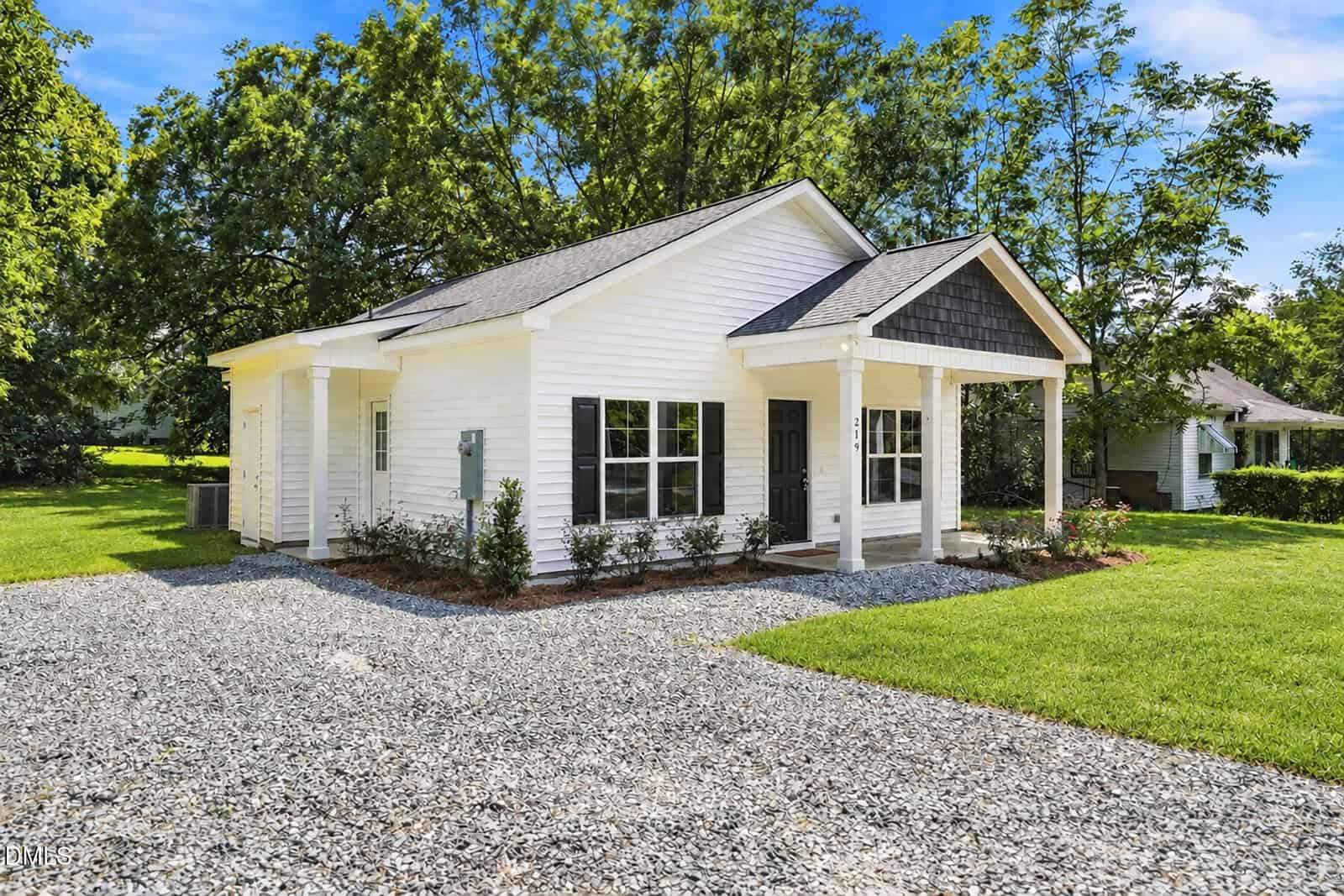 Small white house with a covered front porch, black door, and gravel driveway. Green grass and trees surround the home on a sunny day.