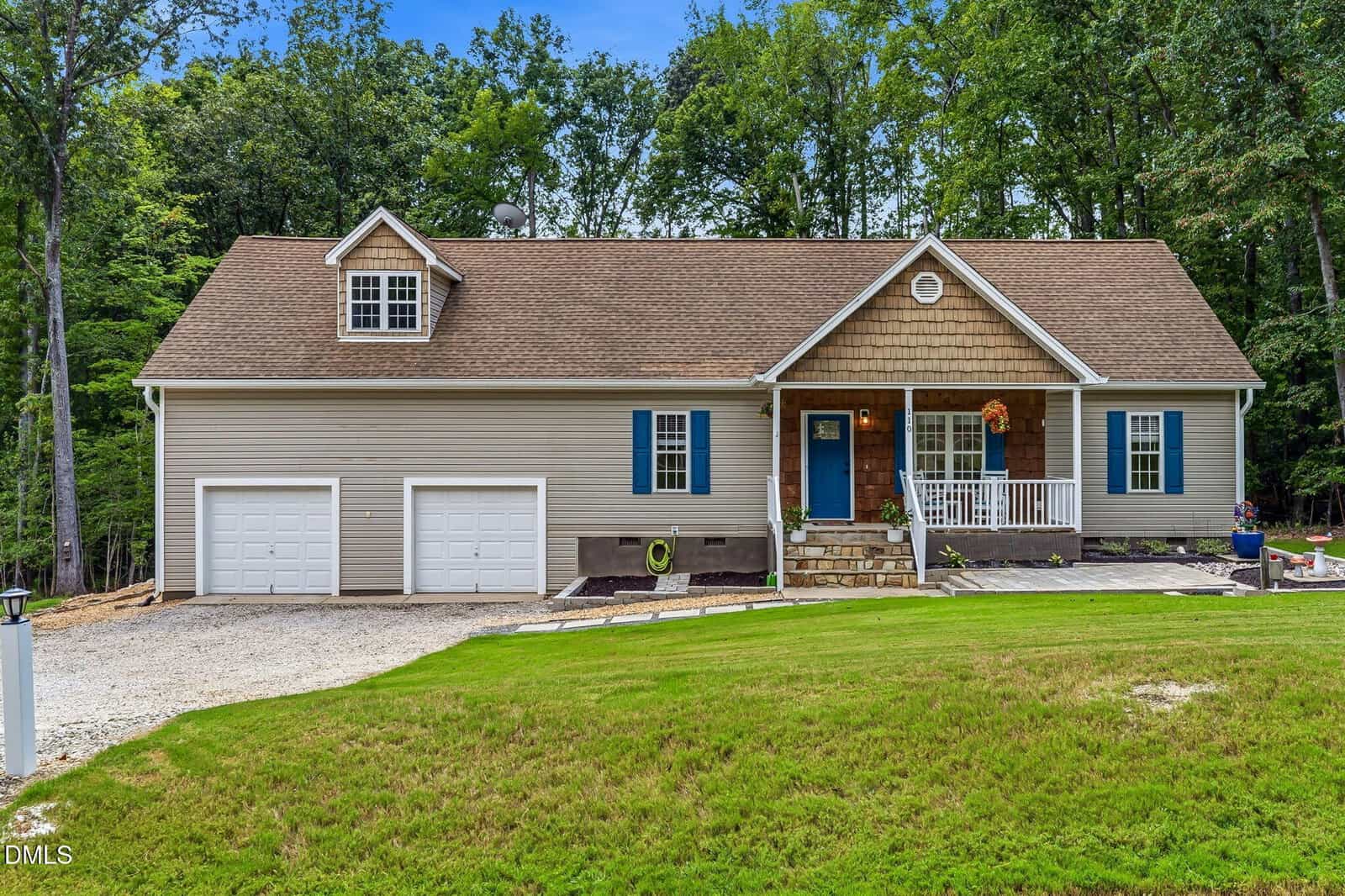 Single-story house with tan siding, front porch, blue door, two-car garage, and a green lawn, surrounded by trees in the background under a blue sky.