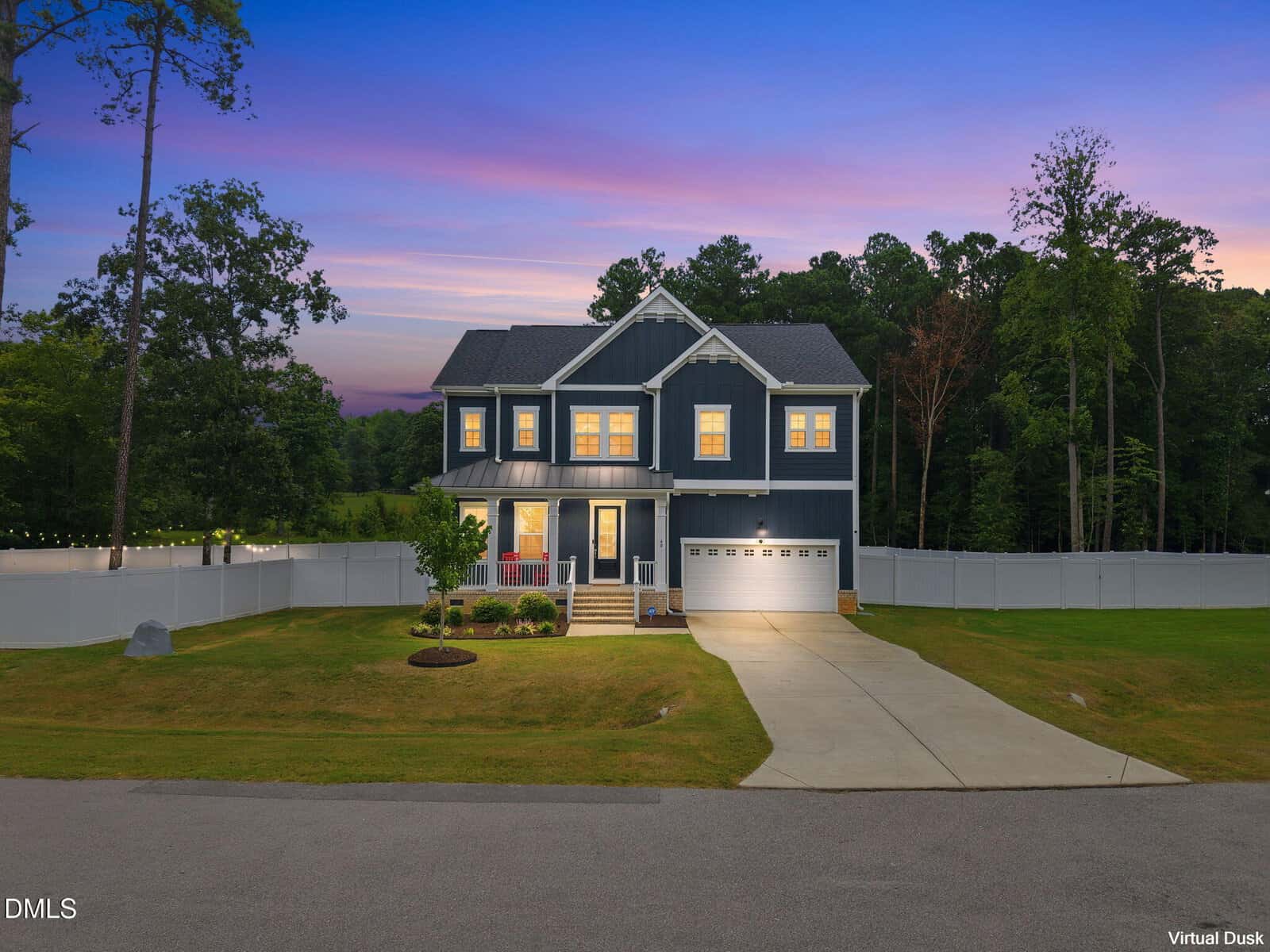 Two-story blue house with white trim, front porch, and attached garage. Exterior lights are on at dusk. Well-kept lawn, surrounding trees, white fence, and a colorful sky in the background.