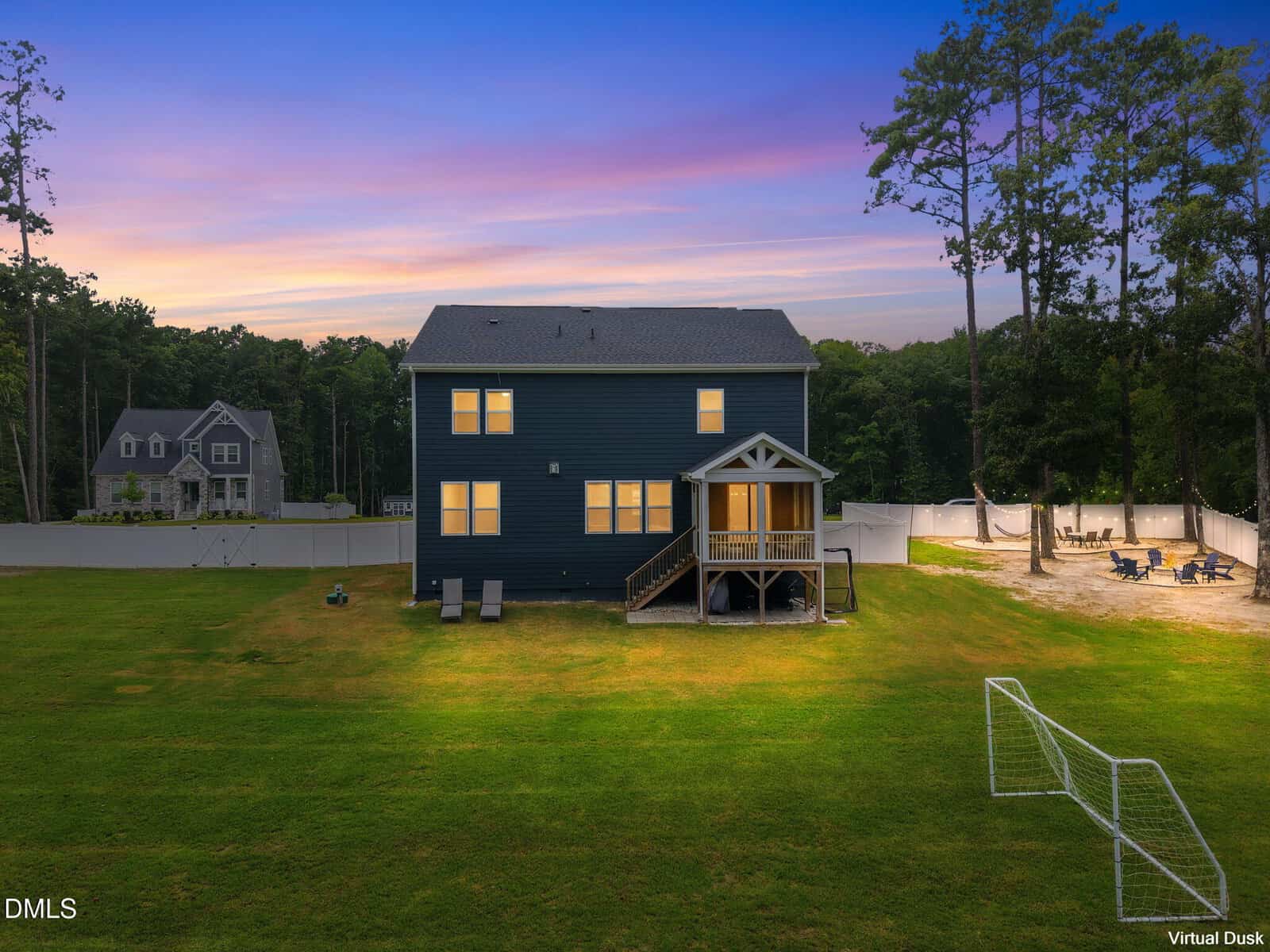 Two-story dark blue house with a small porch, large backyard, soccer net, fire pit area, trees, and a neighboring house. The sky shows a colorful sunset. Virtual dusk effect applied.