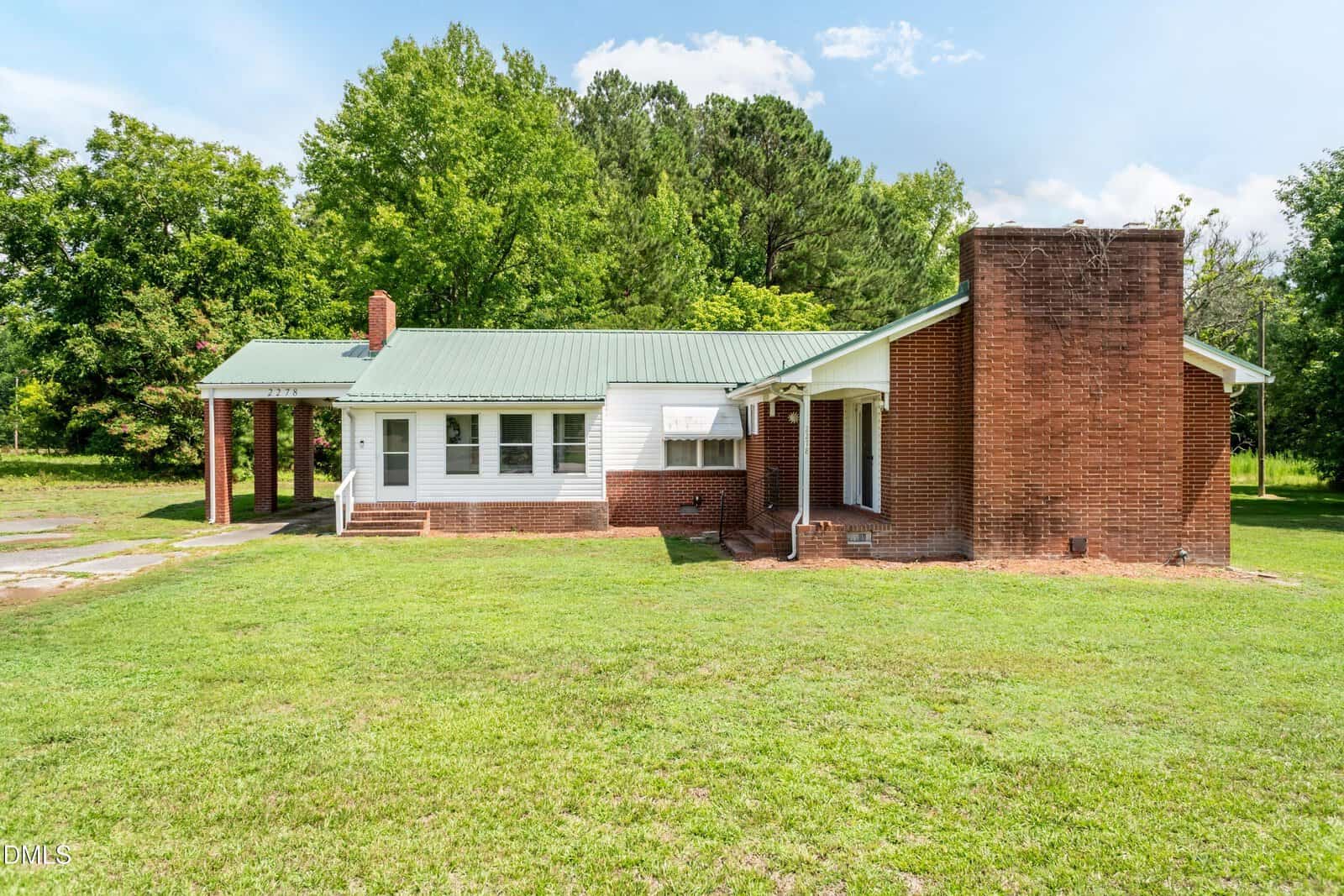 Single-story brick and white house with a green metal roof, large front chimney, and carport, located at 2278 White Level Road in Louisburg. Surrounded by grass and trees on a sunny day, this charming home offers both comfort and curb appeal.