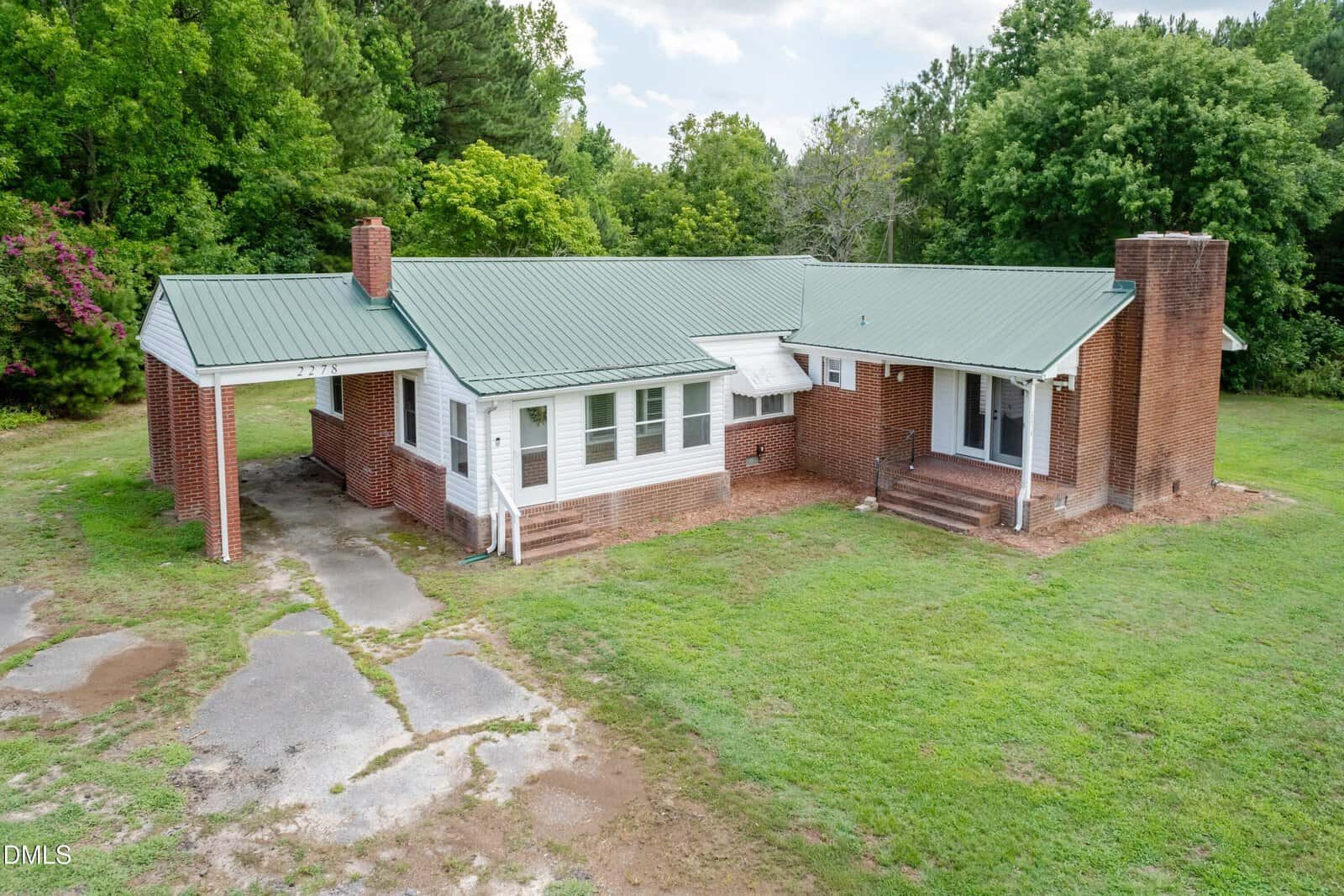 Single-story brick house with green metal roof, covered carport, two chimneys, and front porch. Surrounded by grass and trees, with a partially cracked driveway leading to the carport.