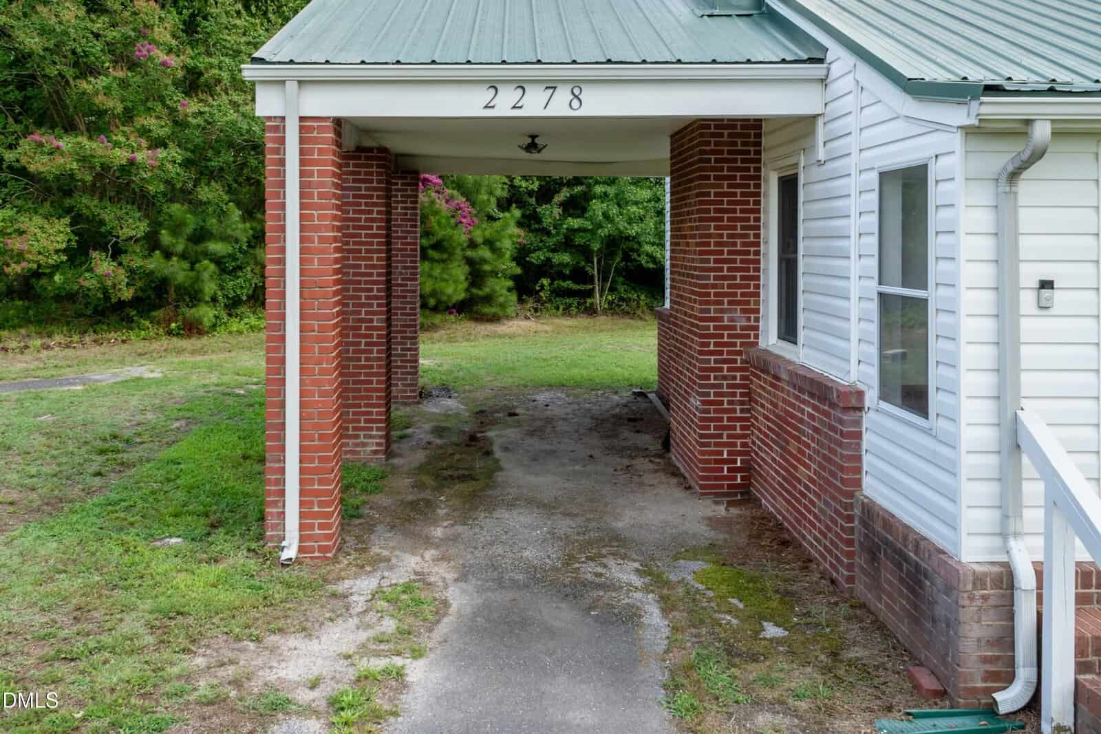 A brick and white siding house with a green metal roof, carport labeled 2278, and a patchy driveway leading into a grassy yard with trees in the background.