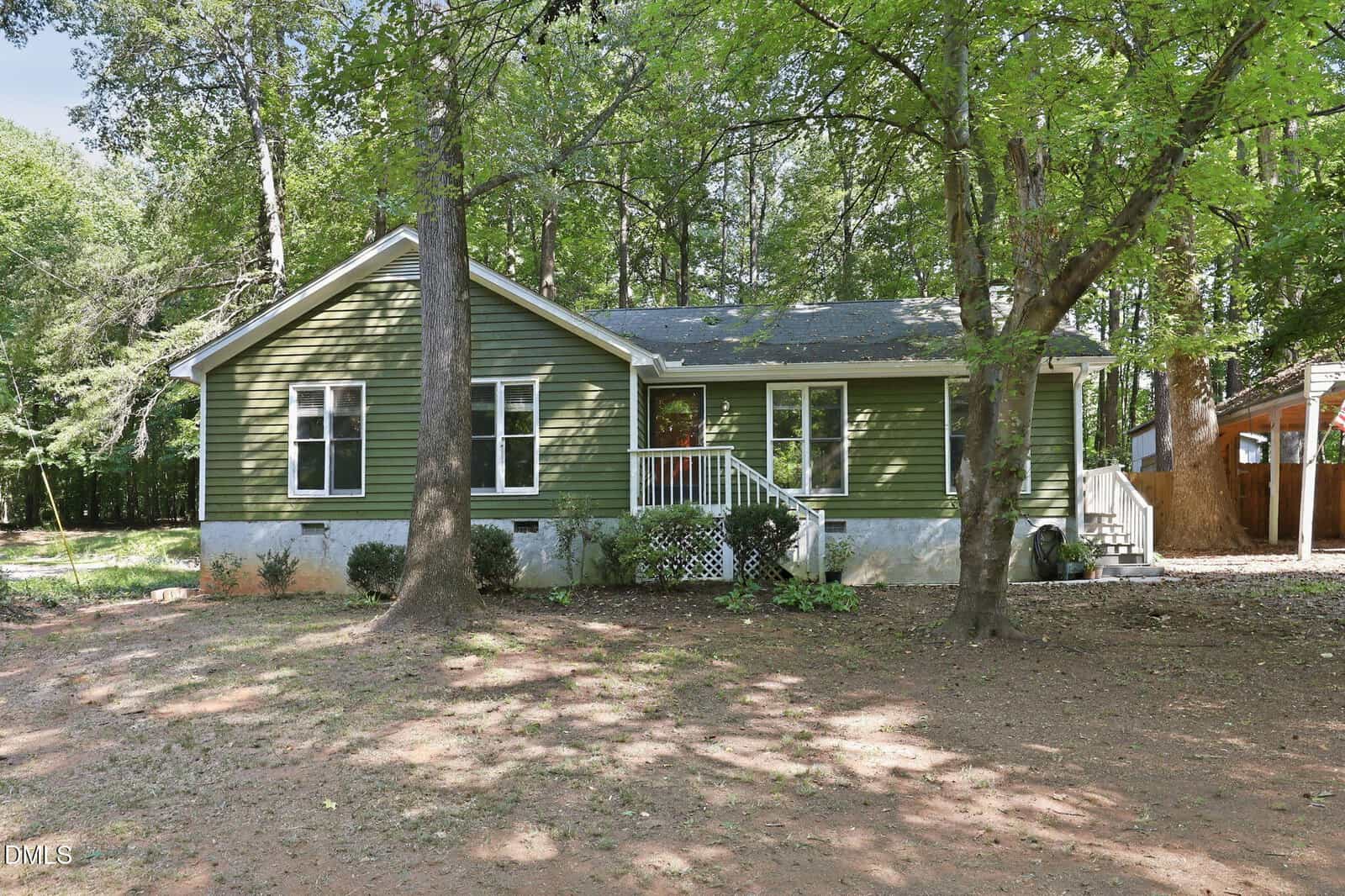 Single-story green house with white trim, a front porch with steps, and a shaded yard surrounded by tall trees on 1016 Holden Road in Youngsville, featuring a dirt ground and wooded background.