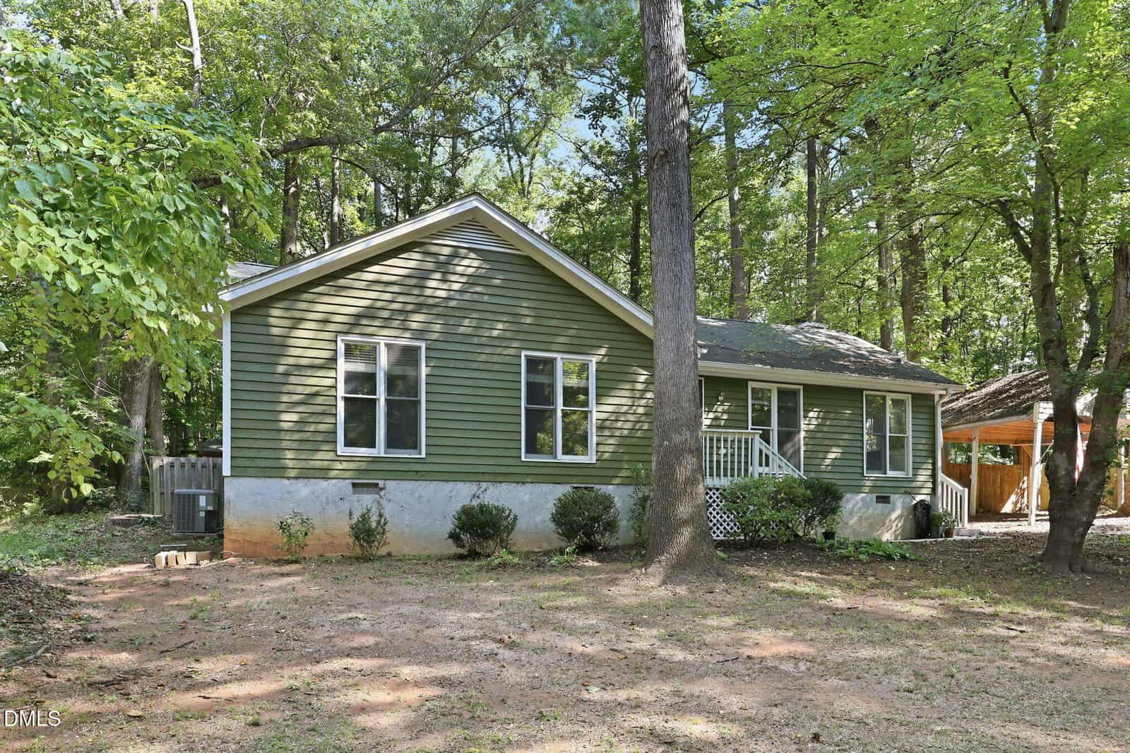 Single-story green house with white trim, a small front porch, carport on the right, surrounded by tall trees and shaded yard with sparse grass.