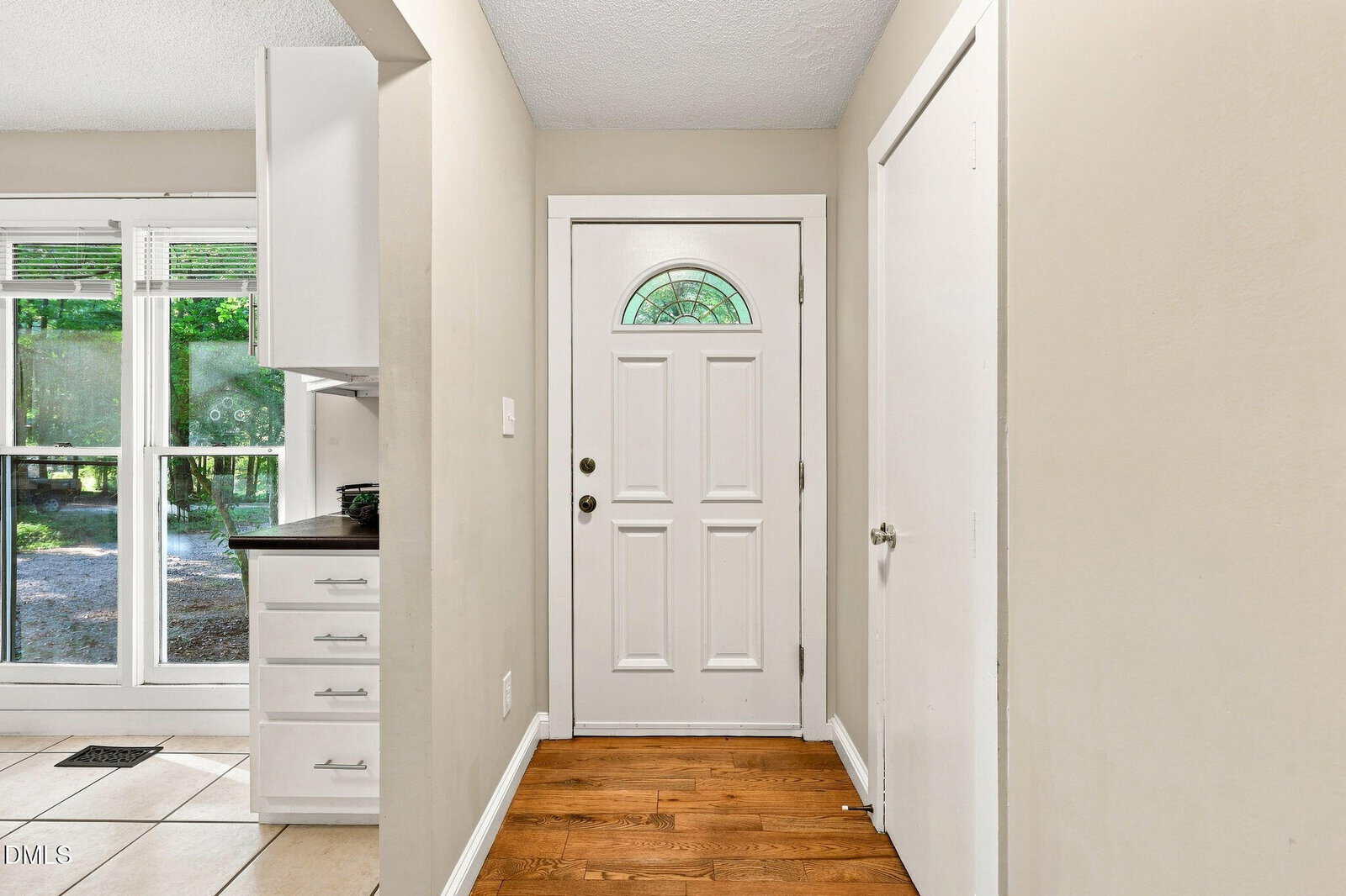 A small entryway with a white front door, a closet door on the right, and hardwood flooring. A partial view of a kitchen with tile floor and cabinets is on the left. Sunlight streams through windows in the background.