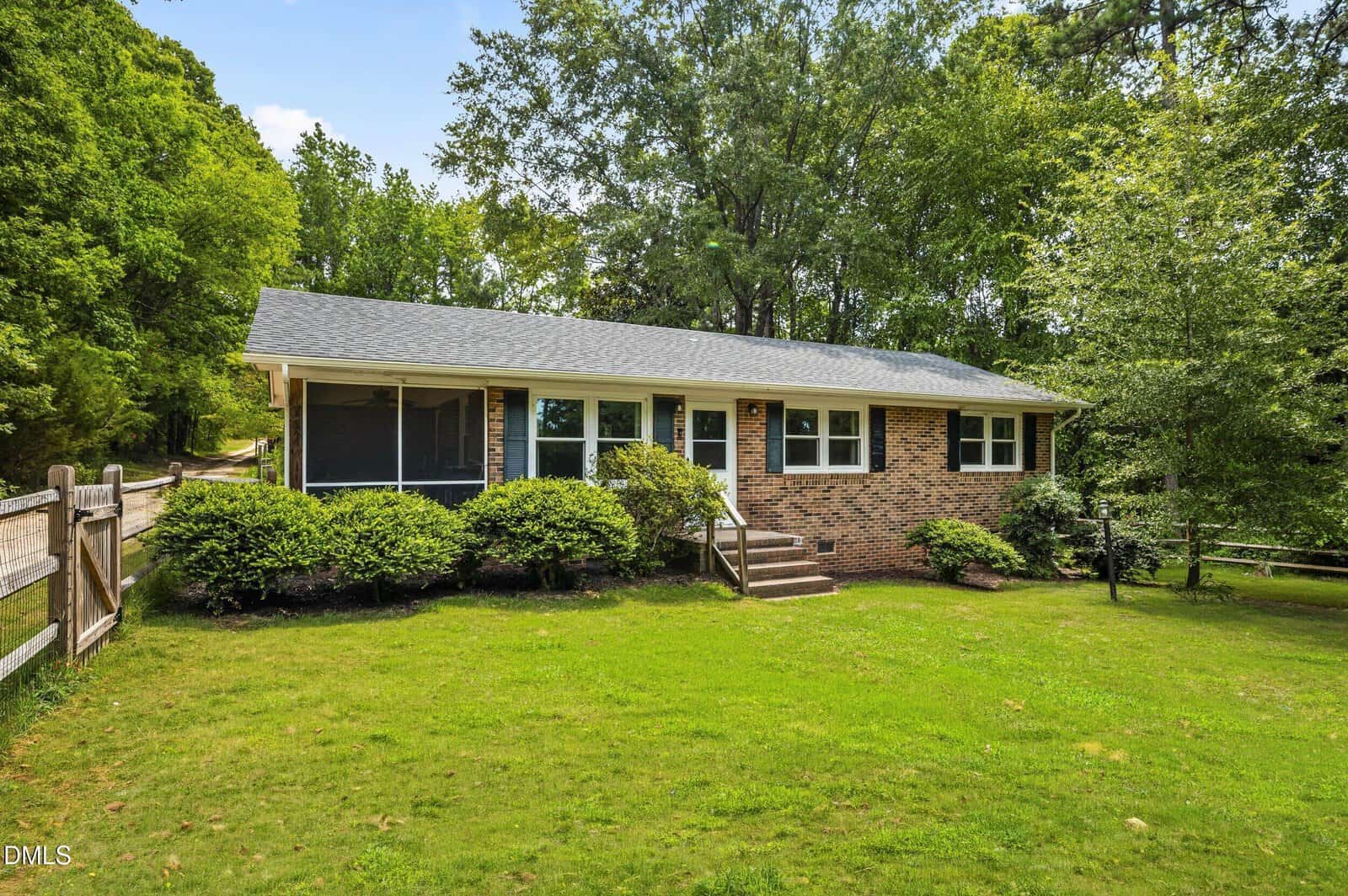 Charming single-story brick house at 382 Greys Mill Road in Louisburg, featuring a screened porch and steps leading to the front door. Surrounded by a lush green lawn, bushes, and mature trees on a sunny day, this home also offers a wooden fence visible on the left side.