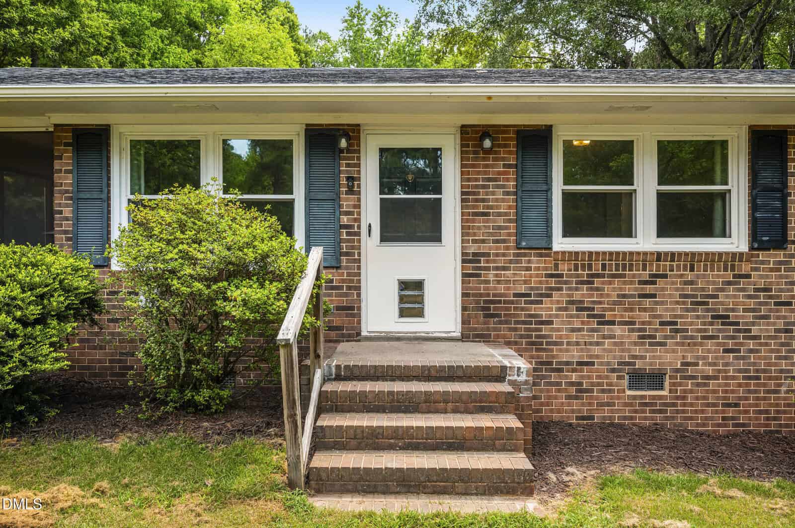 A brick house exterior with a white door, small pet door, blue shutters, and four windows. Bushes and grass are in front, with four brick steps leading to the entrance.