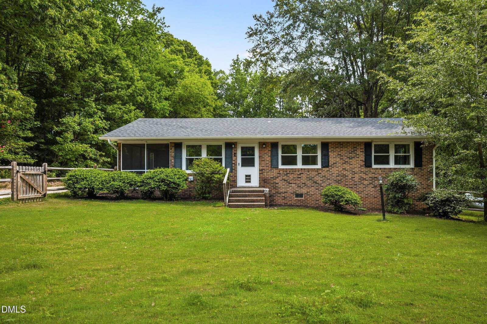 Single-story brick house with dark shutters, a gray roof, and front steps. The home is surrounded by green grass, bushes, and tall trees in a lush, wooded setting.