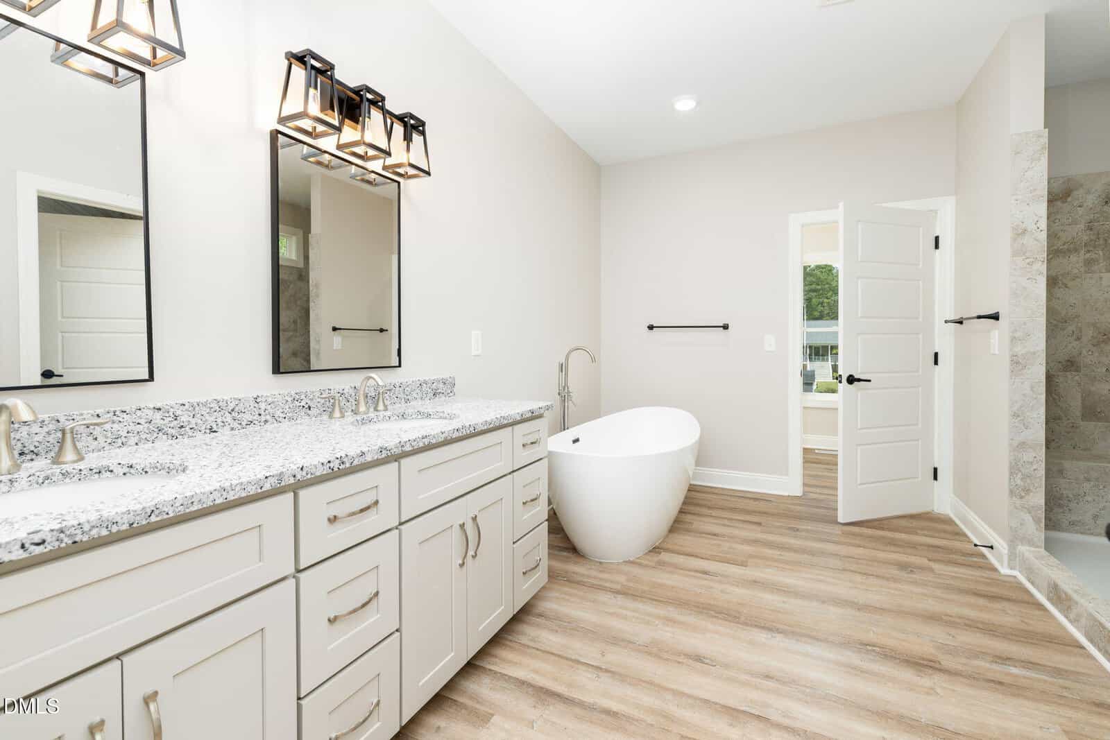 Modern bathroom with double-sink vanity, granite countertop, two mirrors, a freestanding oval bathtub, wood-look flooring, and a walk-in shower. White walls and doors add to the bright, clean appearance.