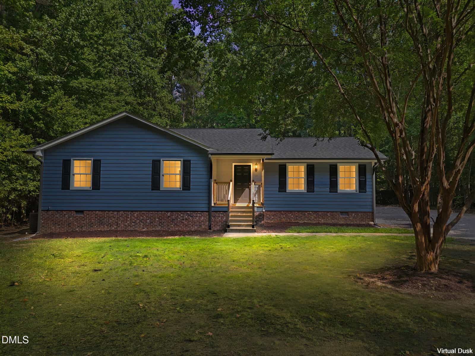 A blue single-story house with a brick foundation is lit warmly from inside at dusk. Trees surround the house, and the front steps lead to a small porch. A patch of lawn is visible in the foreground.