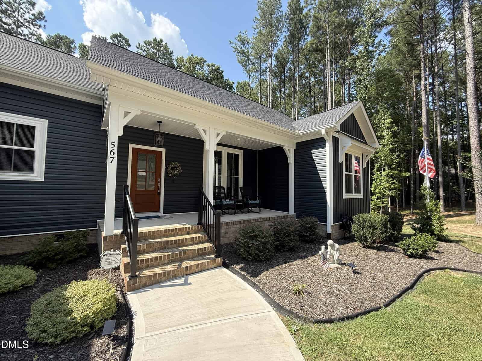 A charming blue house with a front porch, white trim, and a wooden door awaits at 567 Dabney Woods Lane in Henderson. Two rocking chairs sit on the porch near a wreath, while an American flag is proudly displayed. Trees fill the background, and a curved sidewalk leads visitors to the steps.