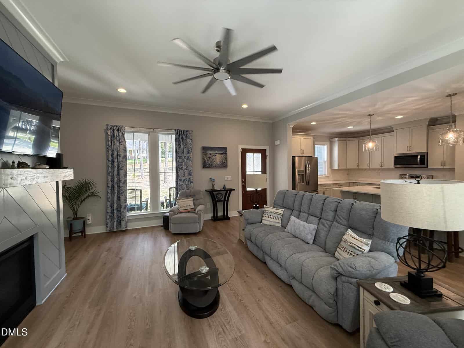 A bright living room with a gray sofa, armchair, round glass coffee table, large TV, ceiling fan, and open kitchen with white cabinets and pendant lights. Light wood floors and a window with blue curtains face the backyard.