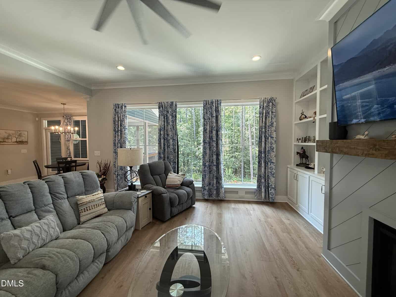 Bright living room with gray sofas, a TV mounted above a fireplace, large windows with blue curtains, shelves with decor, and a view of trees outside. Dining area is visible in the background.