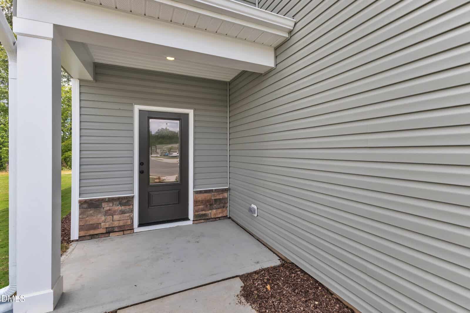 Covered entryway with gray horizontal siding, a black door with a window, brick detail at the base, concrete step, white column, and a view of the street through the door’s glass.