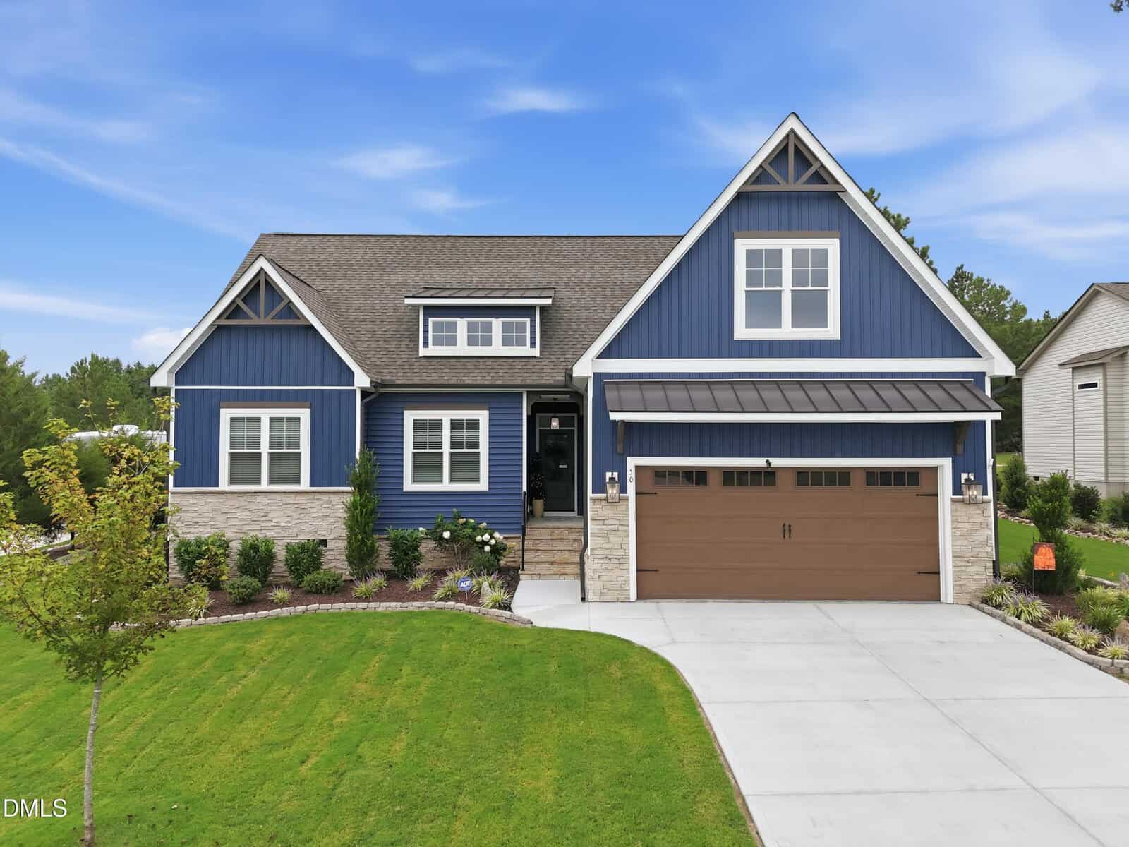 A modern blue house with white trim, stone accents, and a brown garage door sits on a manicured lawn with shrubs and a curved driveway under a partly cloudy blue sky at 50 Basil Court in Franklinton.