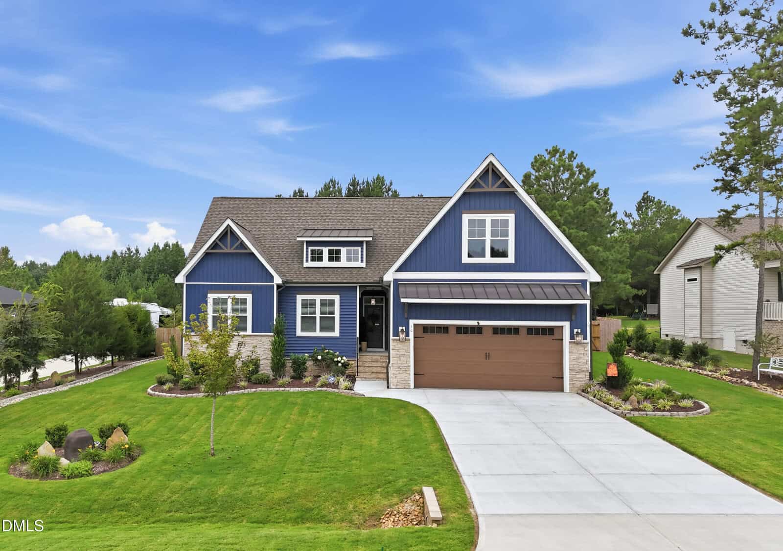 A modern blue house with beige trim, stone accents, and a brown double garage sits on a neatly manicured lawn with trees and a concrete driveway under a bright blue sky.
