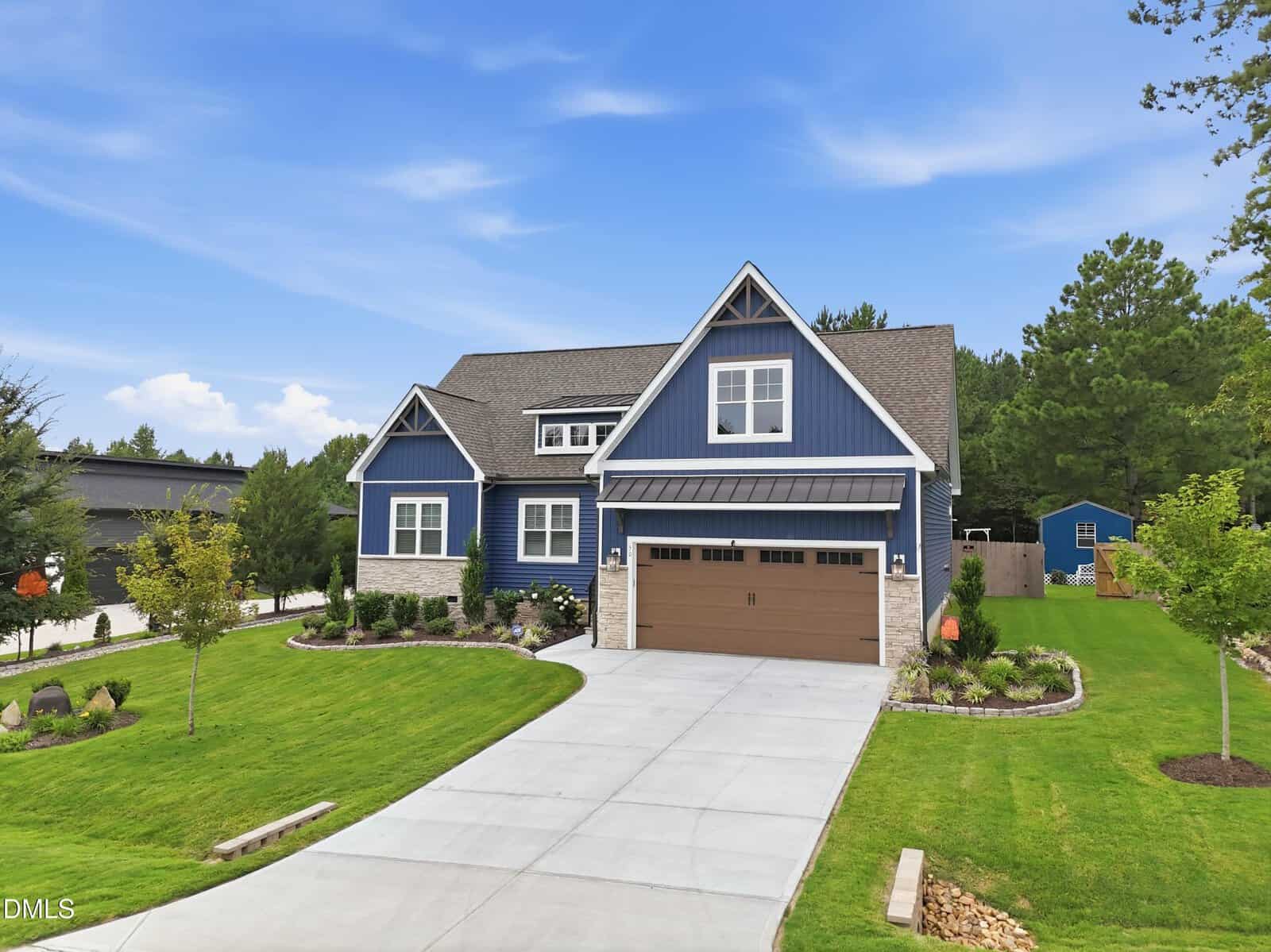 Modern blue house with stone accents, a manicured lawn, and a wide driveway. The yard features landscaped gardens, young trees, and a fenced backyard, set against a backdrop of trees and a blue sky with scattered clouds.