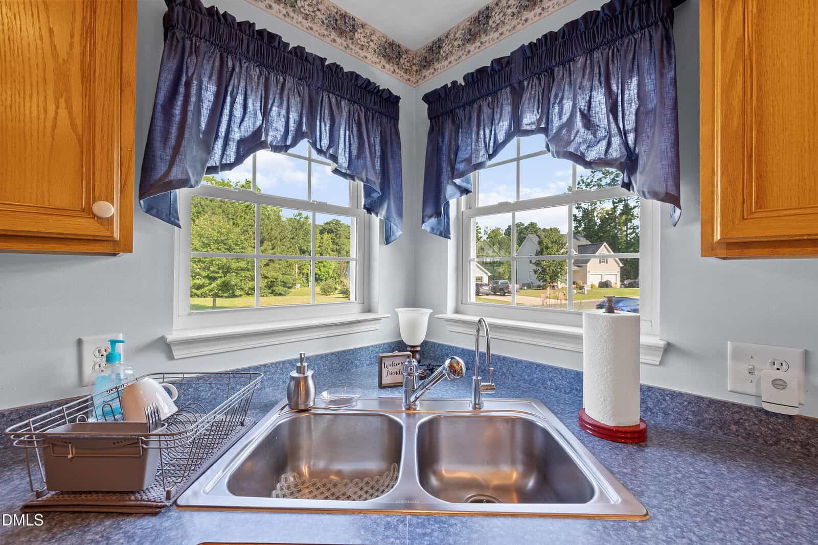 A kitchen sink with a dish rack, soap, and paper towel holder sits under two bright windows with blue curtains, offering a view of a green yard and neighboring houses.