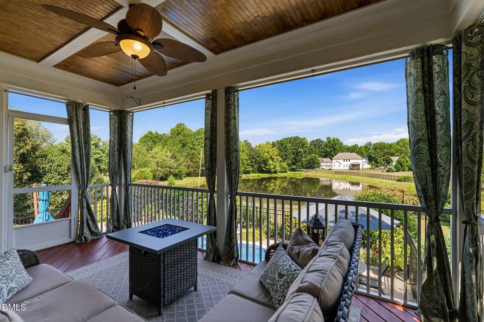 Screened porch with cushioned seating, ceiling fan, and fire table overlooks a pond, trees, and a white house in the distance under a clear blue sky.
